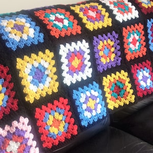 May include: A colorful crocheted blanket made of granny squares in various colors, including red, yellow, blue, green, and purple. The blanket is laid out on a black leather couch.