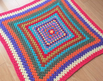 May include: A square crochet blanket featuring a vibrant granny square pattern. The design consists of concentric rows in alternating colors including hot pink, bright orange, turquoise, olive green, purple, and cream, radiating from the center.