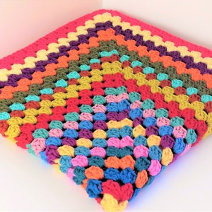 May include: A colourful crocheted blanket with a rainbow pattern. The blanket is made with a variety of different coloured yarns, including red, orange, yellow, green, blue, purple, and pink.