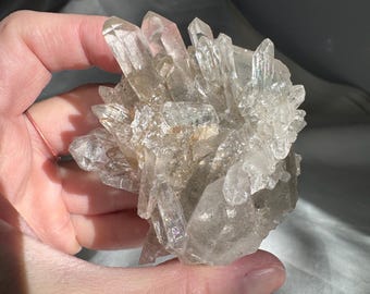 May include: A cluster of clear quartz crystals with a natural, raw appearance. The crystals are clustered together in a rough, organic shape, with some points extending outwards.