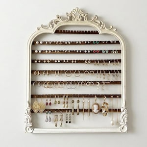 May include: A white, ornate earring organizer with a decorative top and a series of wooden bars. The organizer is filled with various earrings, including studs, hoops, and dangling styles, showcasing a variety of colors and designs.