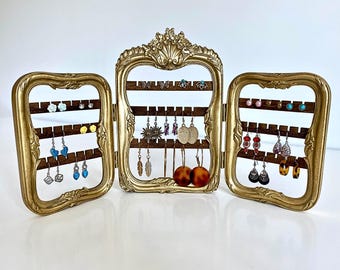 May include: Gold-toned, ornate, three-panel earring holder with a wooden backing. The holder has multiple rows of pegs for displaying earrings. The holder is decorated with intricate details and is a stylish way to organize and display earrings.