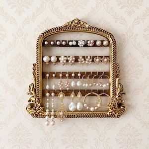May include: A gold-toned, ornate, arched wall-mounted jewelry organizer featuring five horizontal wooden slats with notches for hanging earrings. The display holds a collection of various pearl and gold-toned earrings, including studs, drops, and hoops.