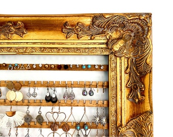 May include: Gold ornate picture frame with a wooden backing and hooks for hanging earrings. The frame is decorated with intricate carvings and has a distressed gold finish.