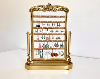 May include: Gold-toned earring stand with a decorative frame and a mirror. The stand has multiple rows of hooks for holding earrings. The stand is on a white surface.
