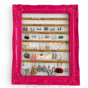 May include: A hot pink framed earring organizer with multiple rows of wooden dowels. The organizer is filled with a variety of earrings in different styles and colors, including dangling, stud, and hoop earrings. The frame has ornate detailing.