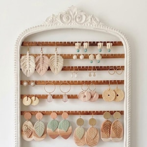 May include: A white, ornate earring display rack filled with various earrings. The earrings include macrame feather designs, gold hoops, and clay leaf shapes in neutral tones. The rack is made of wood and has a decorative white frame.