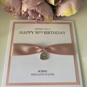 May include: A handmade birthday card with a pink ribbon and a silver charm. The card reads "Wishing you a Happy 50th Birthday" and "Bobbie with lots of love". The card is white with a textured pattern and a pink border.
