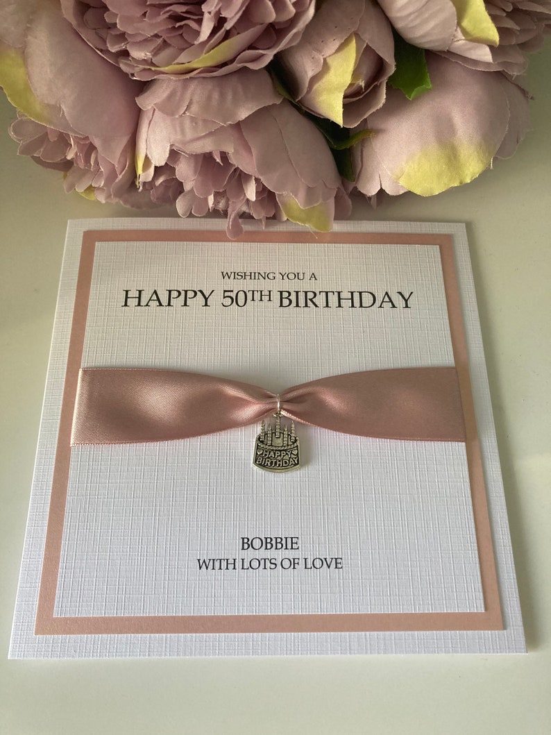May include: A handmade birthday card with a pink ribbon and a silver charm. The card reads "Wishing you a Happy 50th Birthday" and "Bobbie with lots of love". The card is white with a textured pattern and a pink border.