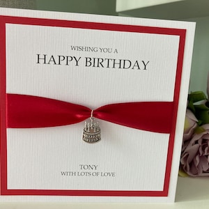 May include: A white birthday card with a red border and a red ribbon. The card says "Wishing you a Happy Birthday" and "Tony with lots of love." A small cake charm hangs from the ribbon.
