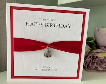 May include: A white birthday card with a red border and a red ribbon. The card says "Wishing you a Happy Birthday" and "Tony with lots of love." A small cake charm hangs from the ribbon.