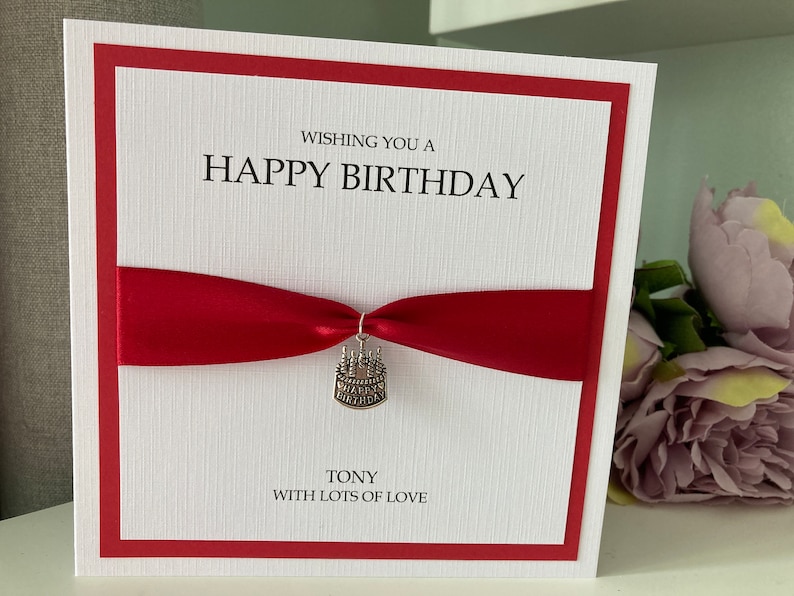 May include: A white birthday card with a red border and a red ribbon. The card says "Wishing you a Happy Birthday" and "Tony with lots of love." A small cake charm hangs from the ribbon.