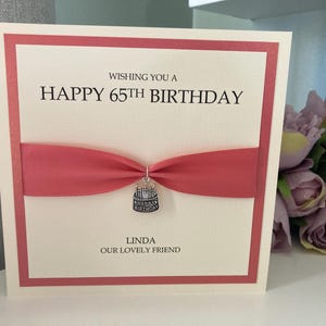 May include: A cream-colored birthday card with a red border and ribbon. The card reads "Wishing you a Happy 65th Birthday" and is personalized with the name "Linda". A small charm shaped like a cake hangs from the ribbon.
