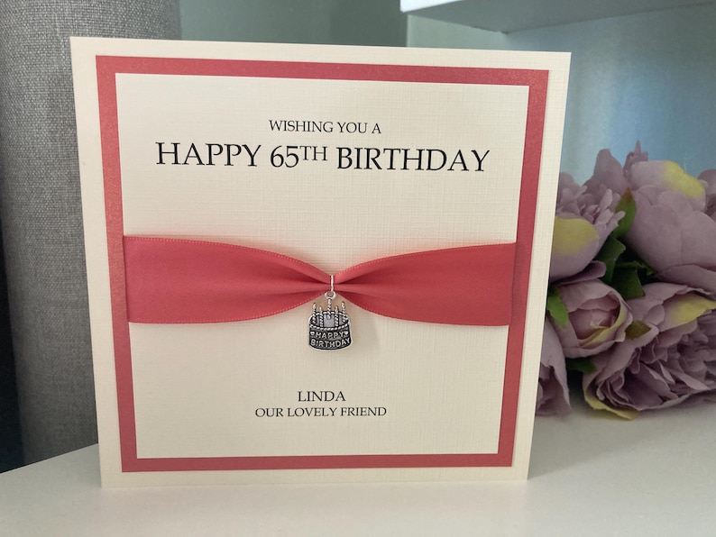 May include: A cream-colored birthday card with a red border and ribbon. The card reads "Wishing you a Happy 65th Birthday" and is personalized with the name "Linda". A small charm shaped like a cake hangs from the ribbon.