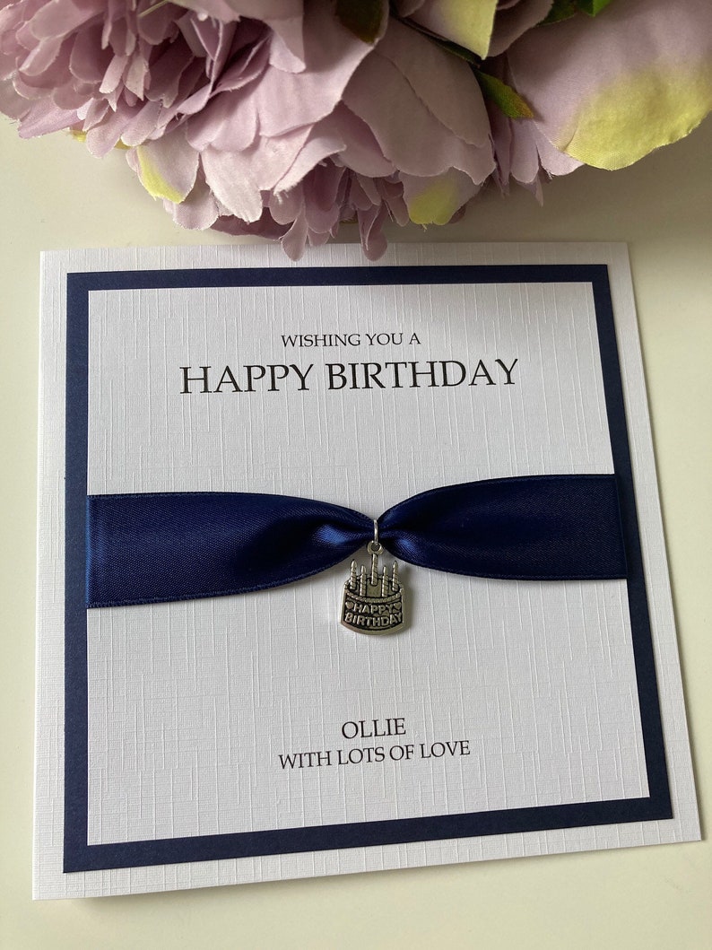 May include: A white birthday card with a navy blue border and a navy blue ribbon. The card reads "Wishing you a Happy Birthday" and "Ollie with lots of love." A small cake charm hangs from the ribbon.