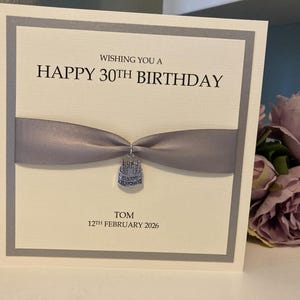 May include: A birthday card with a gray ribbon and a small charm. The card reads "Wishing you a Happy 30th Birthday" with the name "Tom" and the date "12th February 2026" below. The card has a white background with a gray border.