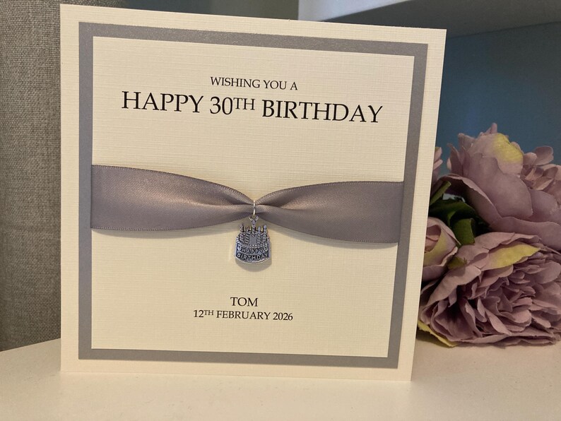 May include: A birthday card with a gray ribbon and a small charm. The card reads "Wishing you a Happy 30th Birthday" with the name "Tom" and the date "12th February 2026" below. The card has a white background with a gray border.