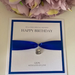 May include: A birthday card with a white textured background, a blue border, and a blue ribbon. The card reads "TO A VERY SPECIAL NEPHEW HAPPY BIRTHDAY" and includes a cake charm. The name "LEON" and "WITH LOTS OF LOVE" are also printed on the card.