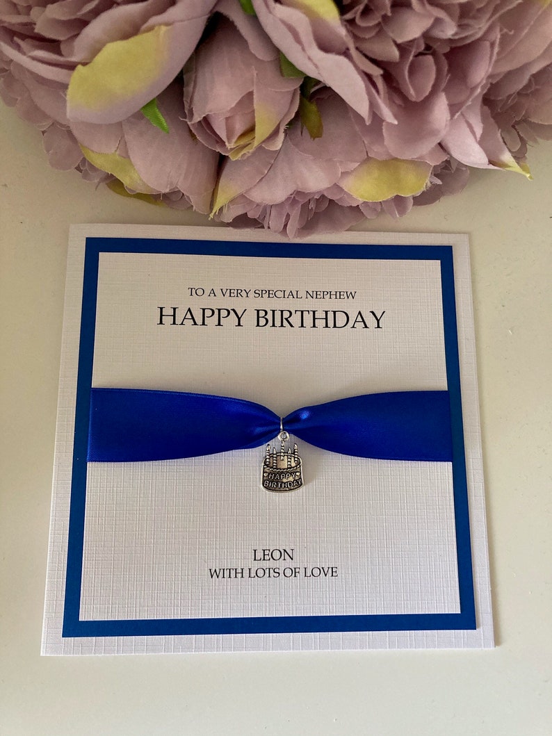 May include: A birthday card with a white textured background, a blue border, and a blue ribbon. The card reads "TO A VERY SPECIAL NEPHEW HAPPY BIRTHDAY" and includes a cake charm. The name "LEON" and "WITH LOTS OF LOVE" are also printed on the card.