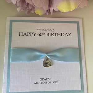 May include: A white birthday card with a light blue border and ribbon. The card reads "WISHING YOU A HAPPY 60th BIRTHDAY" in black text. A small silver charm with "HAPPY BIRTHDAY" is attached to the ribbon. The card is personalized with the name "GRAEME" and the message "WITH LOTS OF LOVE".