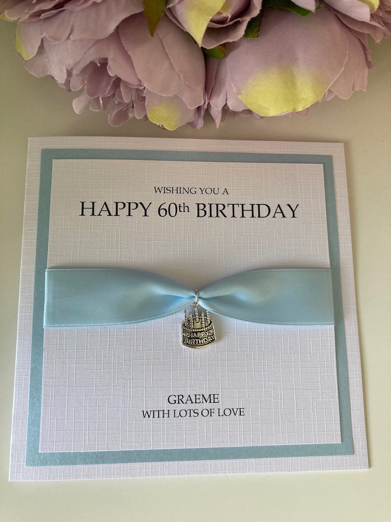May include: A white birthday card with a light blue border and ribbon. The card reads "WISHING YOU A HAPPY 60th BIRTHDAY" in black text. A small silver charm with "HAPPY BIRTHDAY" is attached to the ribbon. The card is personalized with the name "GRAEME" and the message "WITH LOTS OF LOVE".