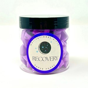 May include: A glass jar filled with purple paper fortune tellers. The jar has a black lid and a label that reads "Motivational Gifts Recovery Paper Phase" with a crescent moon and stars graphic.