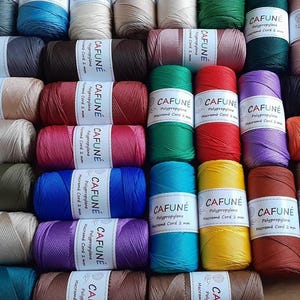 Op de afbeelding: Een verzameling van talloze spoelen van 2 mm polypropyleen macramékoord in een grote verscheidenheid aan effen kleuren. Elke spoel is voorzien van een wit papieren label met de merknaam CAFUNÉ, het materiaal en de diameter in zwarte tekst.
