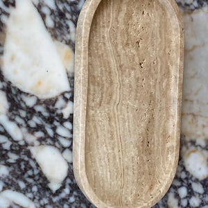May include: An oval-shaped, beige travertine tray with a raised rim. The tray has a natural, porous texture and a light brown color. The background is a marble surface with white and dark gray veining. This tray is suitable for home decor.