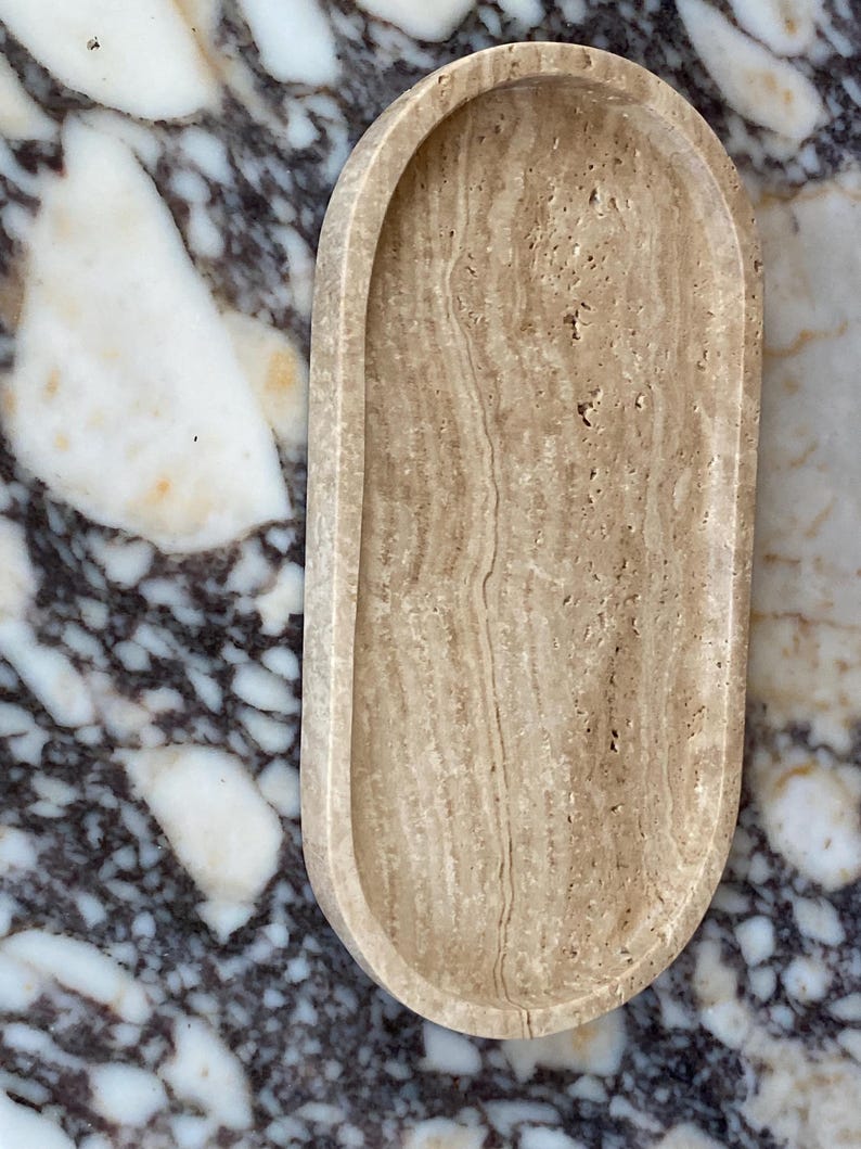 May include: An oval-shaped, beige travertine tray with a raised rim. The tray has a natural, porous texture and a light brown color. The background is a marble surface with white and dark gray veining. This tray is suitable for home decor.