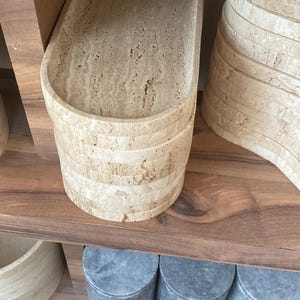 May include: A stack of beige stone trays with a textured surface, displayed on a wooden shelf. The trays have an oval shape and are made of layered stone. Other stone items are also visible.