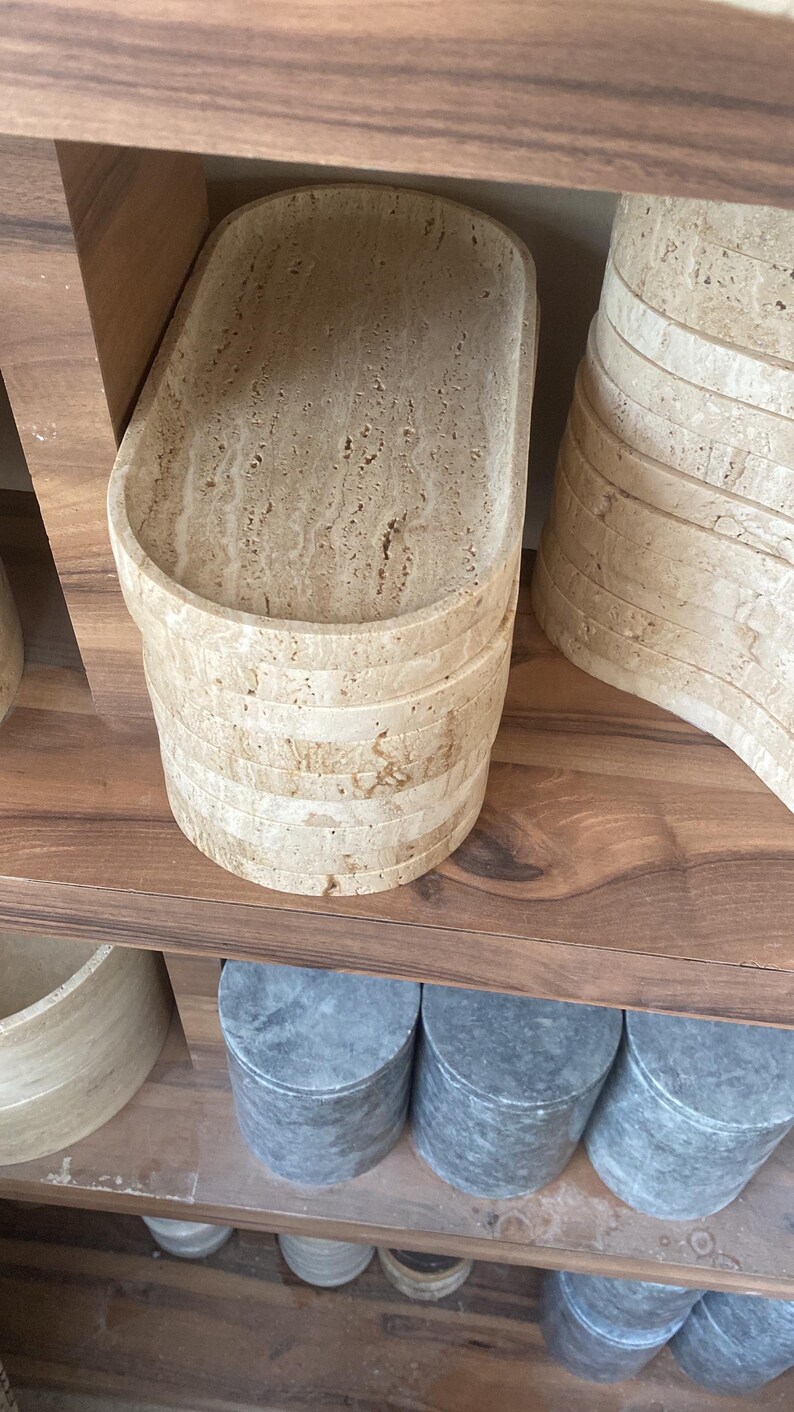 May include: A stack of beige stone trays with a textured surface, displayed on a wooden shelf. The trays have an oval shape and are made of layered stone. Other stone items are also visible.