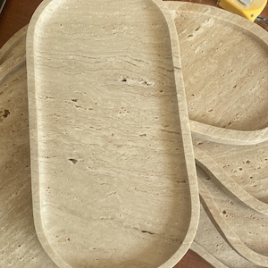 May include: A stack of beige stone trays with a natural, textured appearance. The trays have an oval and circular shape, with a smooth, slightly raised edge. The stone has a marbled effect, with variations in color and pattern.