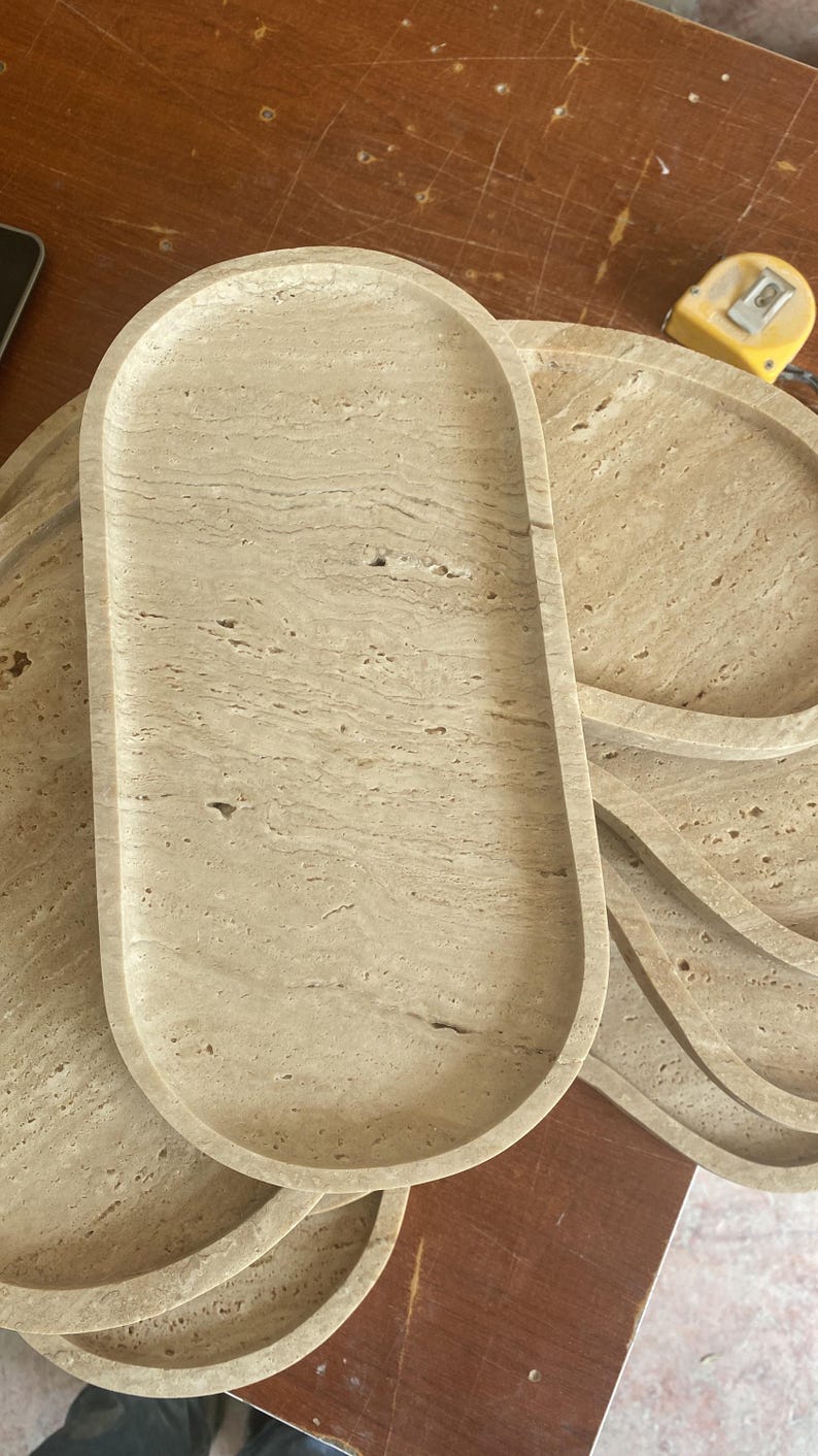 May include: A stack of beige stone trays with a natural, textured appearance. The trays have an oval and circular shape, with a smooth, slightly raised edge. The stone has a marbled effect, with variations in color and pattern.
