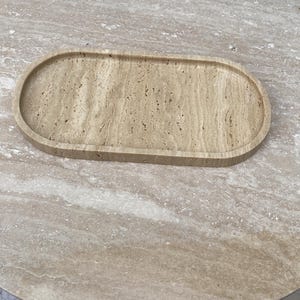 May include: A beige, oval-shaped tray made of stone, resting on a matching stone table. The tray has a slightly raised edge and a textured surface, showcasing natural patterns. The table has a round shape.