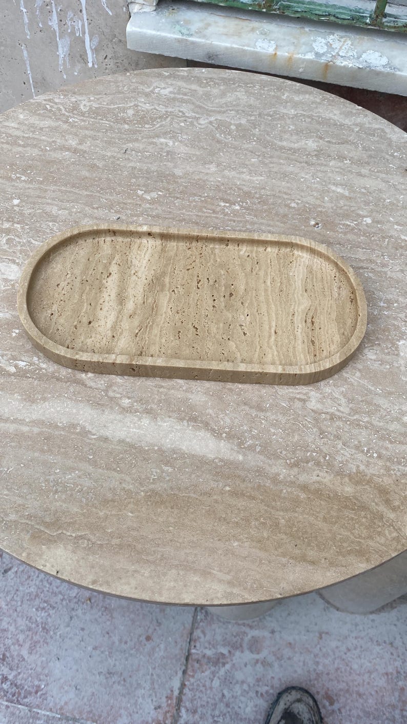 May include: A beige, oval-shaped tray made of stone, resting on a matching stone table. The tray has a slightly raised edge and a textured surface, showcasing natural patterns. The table has a round shape.