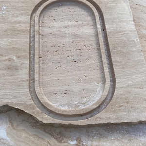 May include: A rectangular piece of beige stone with an oval-shaped recessed area carved into its surface. The stone has a natural, textured appearance with variations in color and pattern, suggesting a natural material.