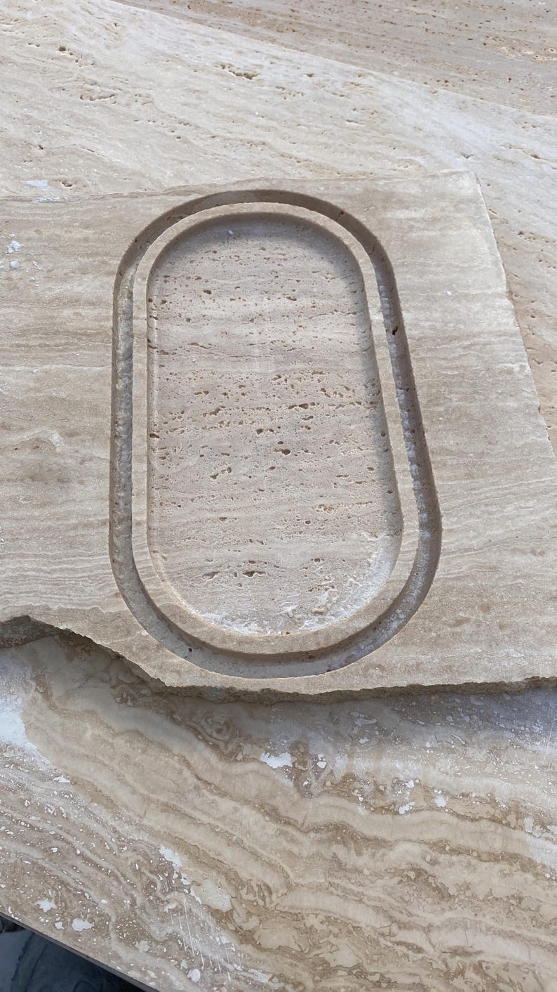 May include: A rectangular piece of beige stone with an oval-shaped recessed area carved into its surface. The stone has a natural, textured appearance with variations in color and pattern, suggesting a natural material.