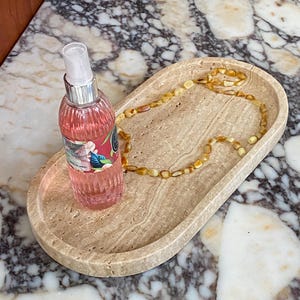 May include: An oval-shaped, beige stone tray holds a pink spray bottle and a beaded amber necklace. The bottle has a silver cap and a label with floral designs. The necklace is made of small, rounded amber beads.