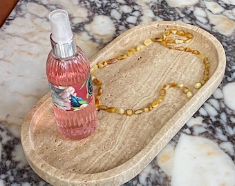 May include: An oval-shaped, beige stone tray holds a pink spray bottle and a beaded amber necklace. The bottle has a silver cap and a label with floral designs. The necklace is made of small, rounded amber beads.