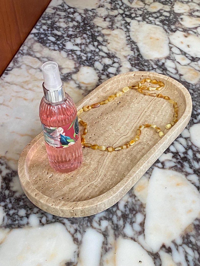 May include: An oval-shaped, beige stone tray holds a pink spray bottle and a beaded amber necklace. The bottle has a silver cap and a label with floral designs. The necklace is made of small, rounded amber beads.