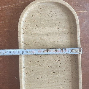 May include: A light beige, oval-shaped tray made of stone. The tray is approximately 10 inches long. The surface has a natural, textured appearance with small holes and variations in color. The tray is on a wooden surface.