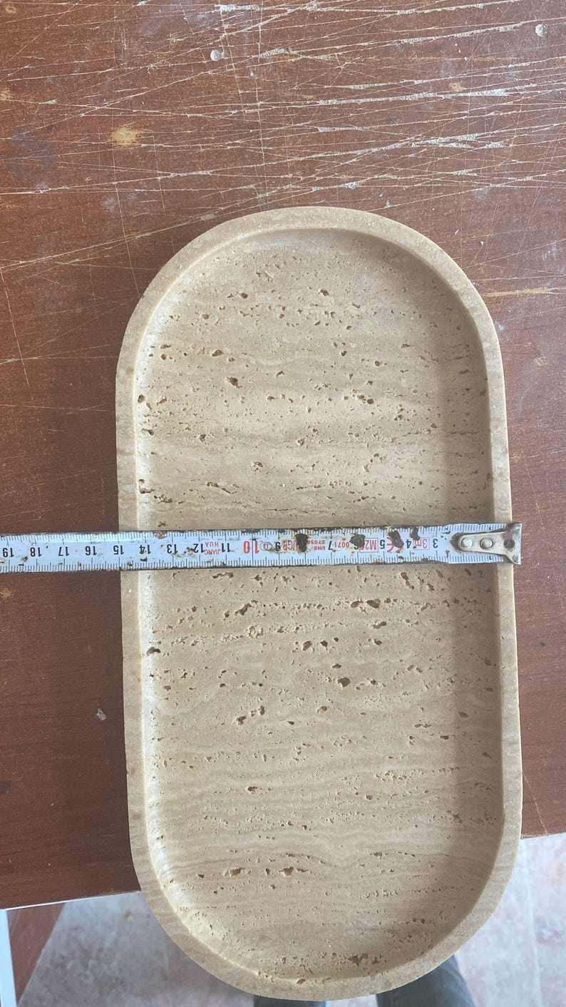 May include: A light beige, oval-shaped tray made of stone. The tray is approximately 10 inches long. The surface has a natural, textured appearance with small holes and variations in color. The tray is on a wooden surface.
