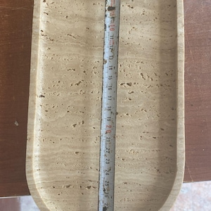 May include: A beige, oval-shaped tray with a textured surface. The tray is approximately 12 inches long. A metal measuring tape is placed on top of the tray, indicating its length. The tray is made of stone.