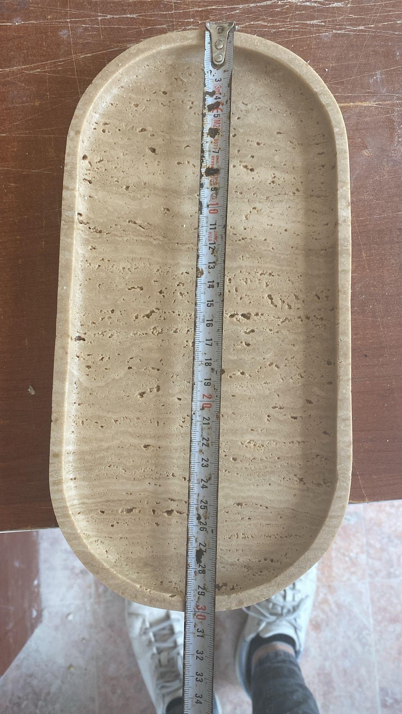 May include: A beige, oval-shaped tray with a textured surface. The tray is approximately 12 inches long. A metal measuring tape is placed on top of the tray, indicating its length. The tray is made of stone.