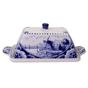 Può includere: Un porta burro in ceramica bianca con un design Delft blu e bianco che presenta un mulino a vento e una scena di canale. Il coperchio ha un pomo in cima.
