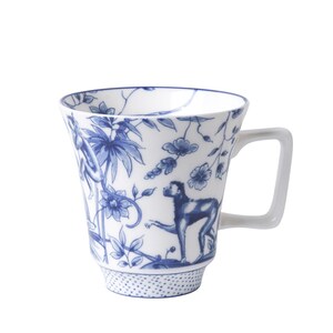 Könnte beinhalten: Eine weiße Keramiktasse mit einem blauen und weißen Blumen- und Affenmuster. Die Tasse hat eine ausgestellte Form und einen Henkel.