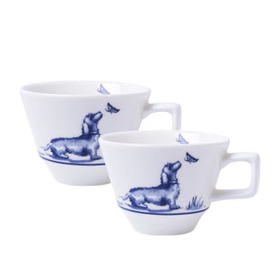 Op de afbeelding: Twee witte keramische mokken met een blauw teckel- en vlinderontwerp. De mokken hebben een vierkant handvat en een blauwe streep aan de basis. De hond kijkt omhoog naar een vlinder.