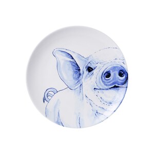 Peut inclure: Une assiette en céramique blanche avec une illustration bleue et blanche d'un visage de cochon. Le cochon regarde vers la droite et a une expression espiègle.