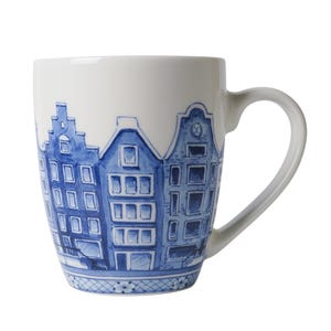Könnte beinhalten: Weißer Keramikbecher mit einer blau aquarellierten Illustration von Grachtenhäusern. Der Becher hat einen gebogenen Henkel und einen dekorativen Rand am Boden. Das Design erinnert an niederländische Architektur.