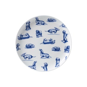 Op de afbeelding: Een wit keramiek bord met een blauw en wit patroon van teckels in verschillende poses. Het bord is rond en heeft een licht verhoogde rand.